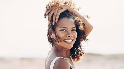 A smiling woman with curly brown hair looks over her shoulder, gently touching her hair with one hand. She wears a white top against a bright, softly blurred background